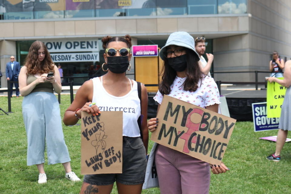 Two women at an outdoor protest holding signs that read "Keep Your Laws Off My Body" and "My Body My Choice." Other people are visible in the background