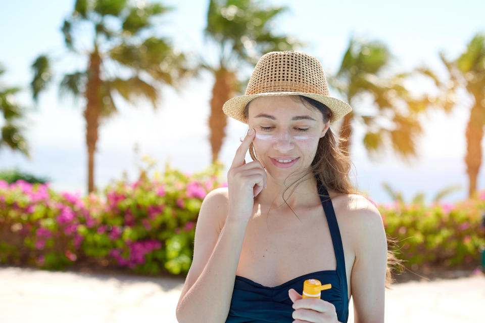 A young woman who is wearing a hat and is smiling in a swimsuit smeared face with sun protection cream 