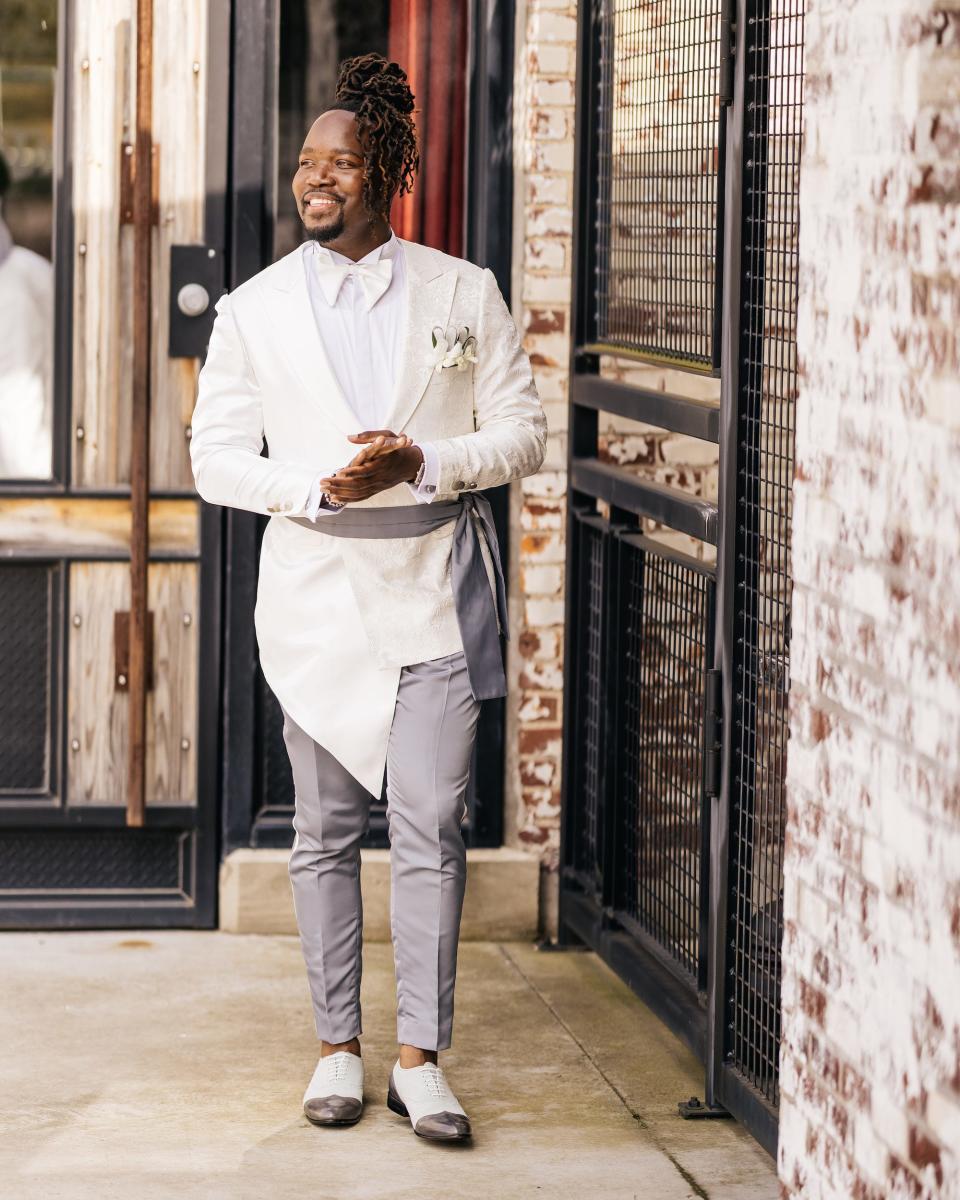 A groom stands in front of a glass door in his wedding attire.