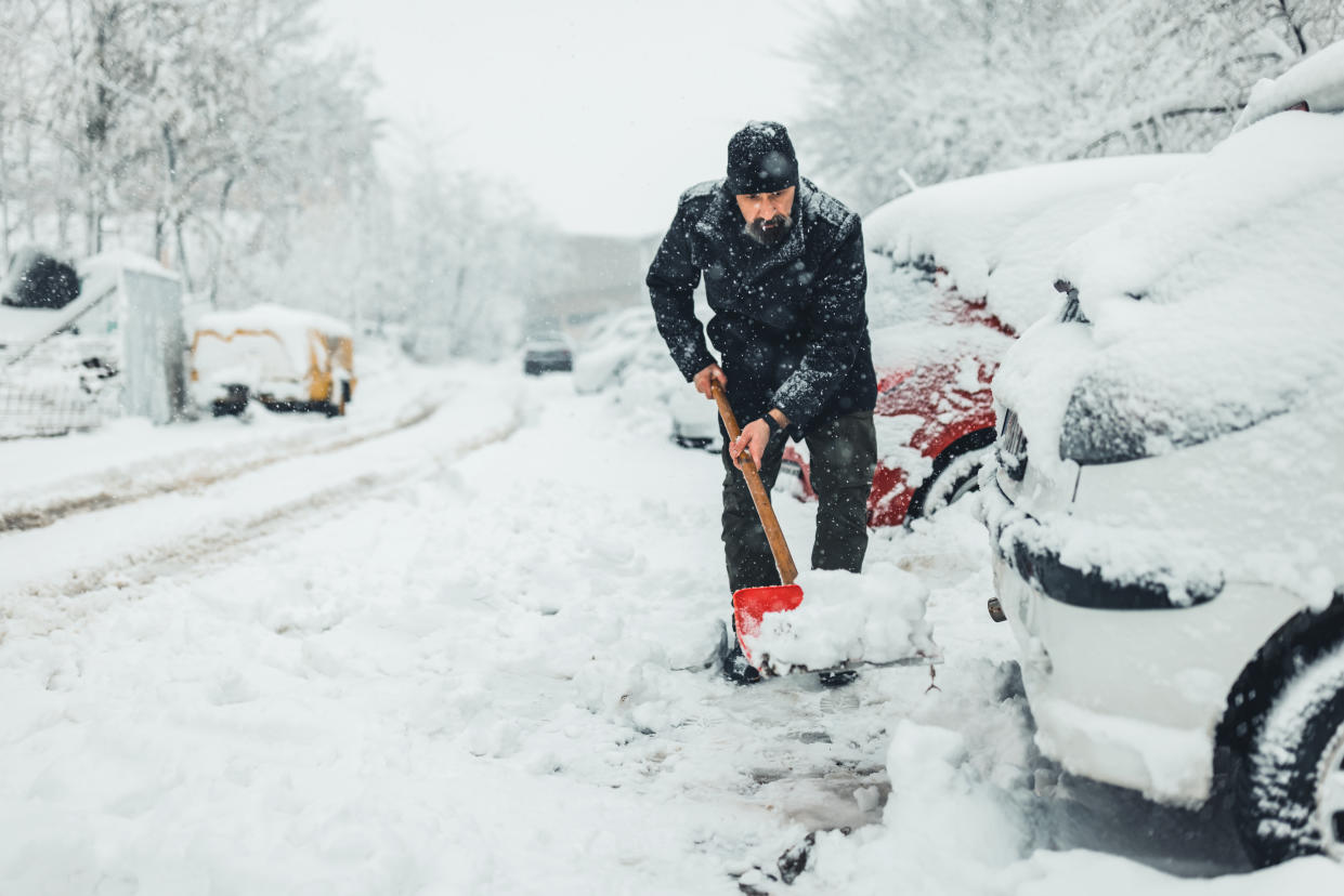 Mature man, male in winter coat cleaning, shoveling driveway, street from covered falling, snow in heavy snowing snowstorm with shovel, residential houses, cars parked on road