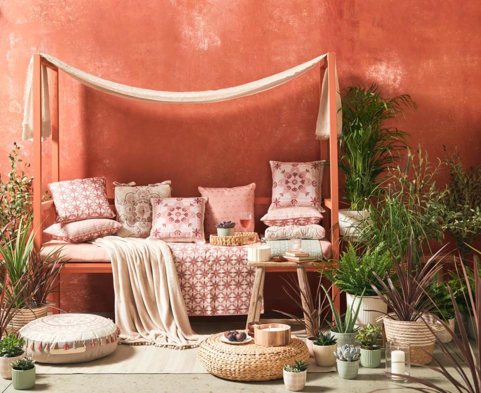 a bench draped with a linen fabric and filled with pillows and throws surround by potted plants against a plaster coral wall