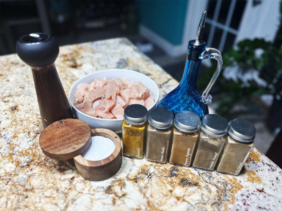 A cup of salt, a pepper grinder, a white bowl of raw chicken pieces, an olive-oil dispenser, and several spices lined up on a marble kitchen counter