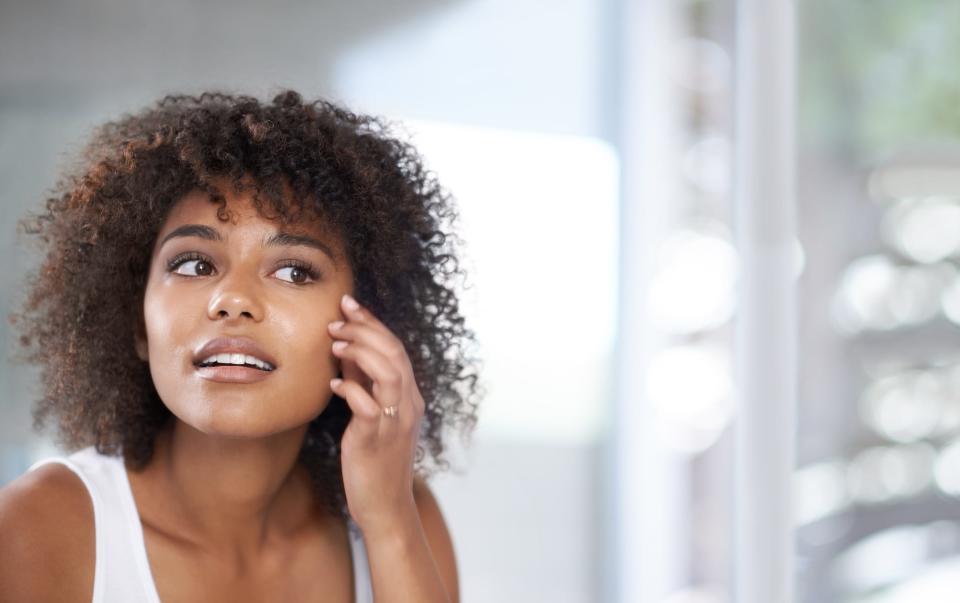 A young woman with dark skin and curly hair looking in a mirror and checking out her healthy skin