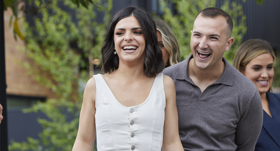 Steph and Gian stand side by side laughing with Eliza in the background and green foliage behind them. 
