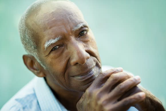 A smiling elderly man resting his chin on his interlocked hands.