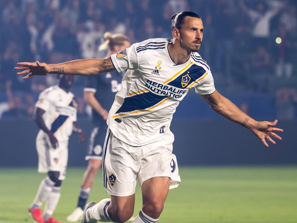 Soccer player Zlatan Ibrahimović wears a white jersey and shorts on the soccer field and spreads his arms wide