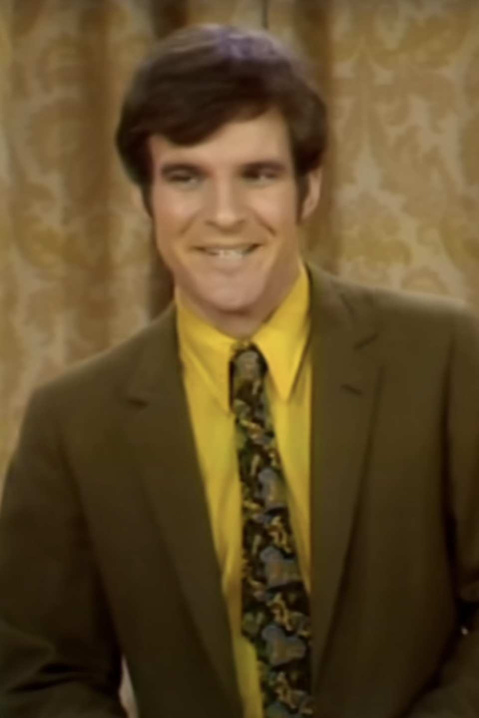 A smiling, young Steve Martin, wearing a brown suit jacket, a yellow shirt, and a patterned tie, stands in front of a curtain