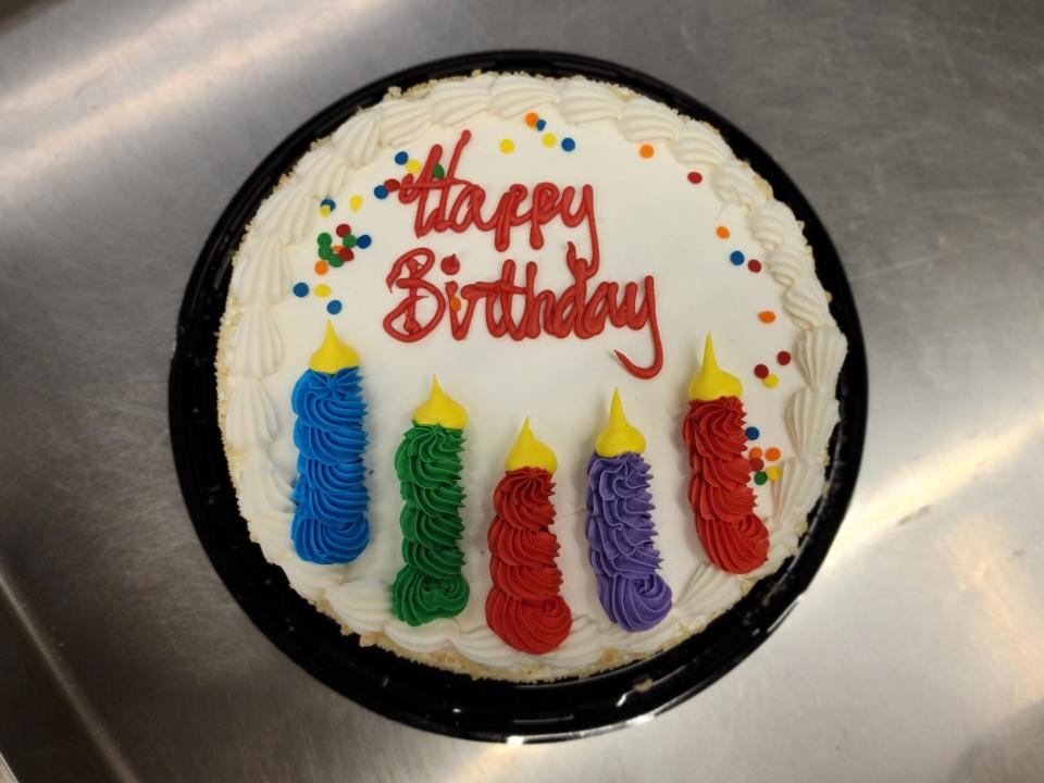 Bird's-eye view of a cake with white frosting and colorful icing in the shape of candles and a red "Happy Birthday" message. The cake is also covered in rainbow sprinkles