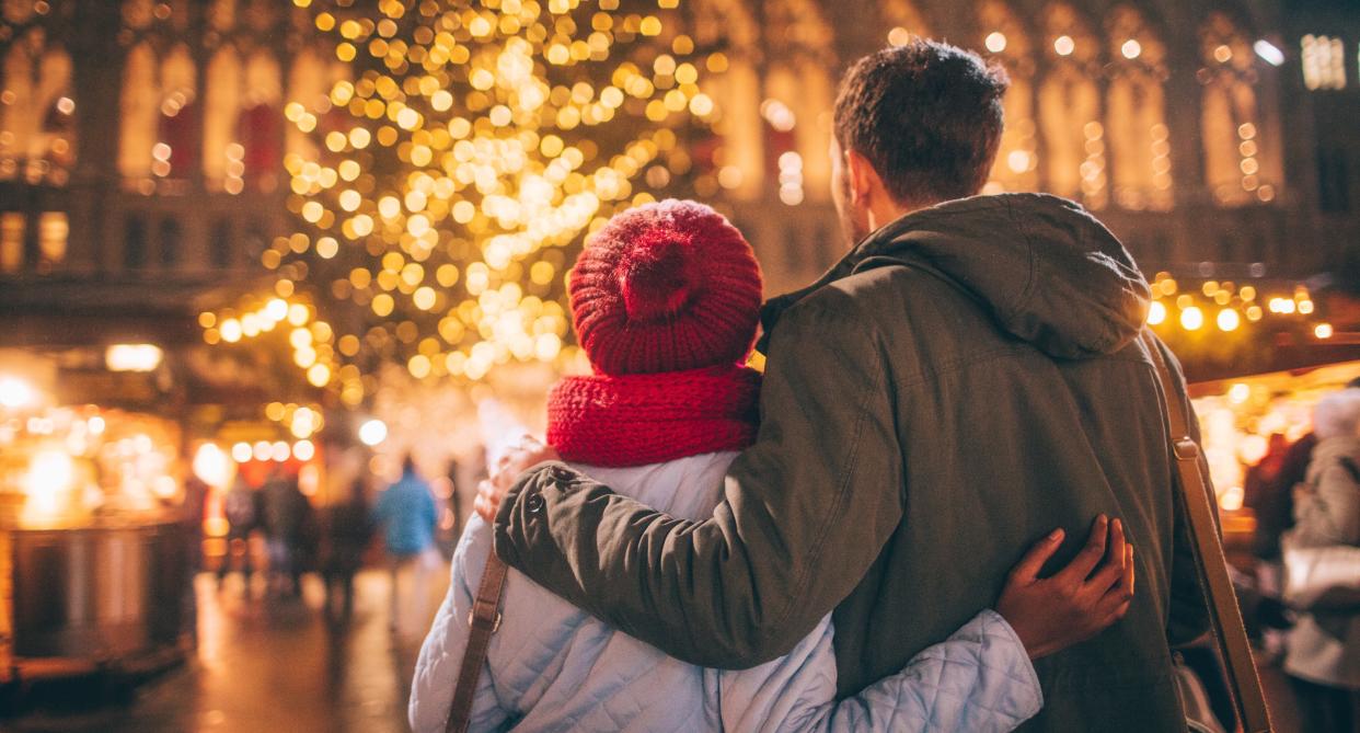 Man and woman embrace in winter clothes as they stand outside in front of a Christmas tree. (Getty Images)