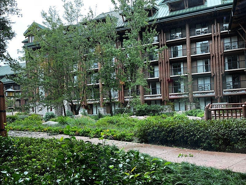 Wilderness lodge, with several balconies and log details, at Disney World. Trees and bushes a side walk with fallen branches and leaves are in the foreground