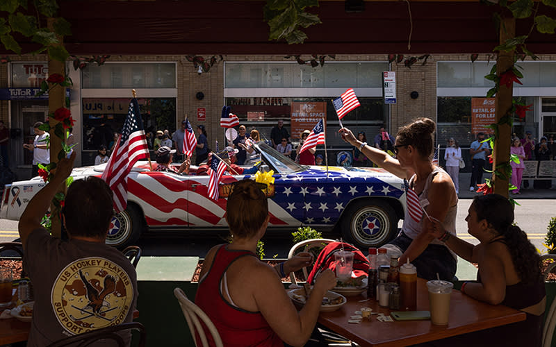People, seated at shaded restaurant patio, wave American flags as participants drive past during Brooklyn's 155th Memorial Day Parade