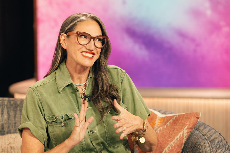 A woman with long wavy hair and glasses wearing a casual button-down shirt is smiling and gesturing while sitting on a sofa against a colorful background.