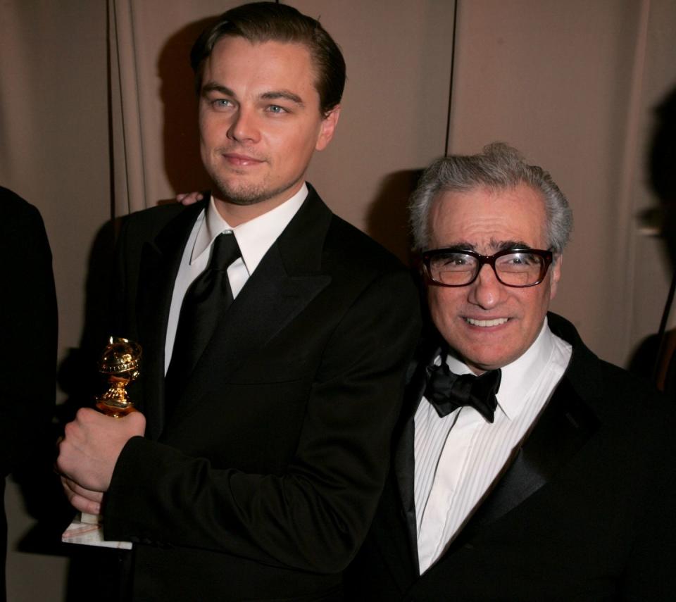 leonardo dicaprio stands next to martin scorsese who has an arm on dicaprios should, dicaprio grips a golden globe statue with both hands and looks to the left of the camera, scorsese smiles and looks to the right of the camera, dicaprio wears a black suit and tie with a white collared shirt, scorsese wears a black tuxedo with a white shirt