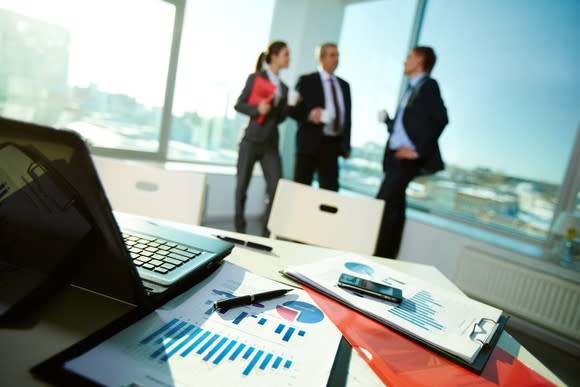 Three businesspeople talk in an office with a computer and papers on a table in the foreground.