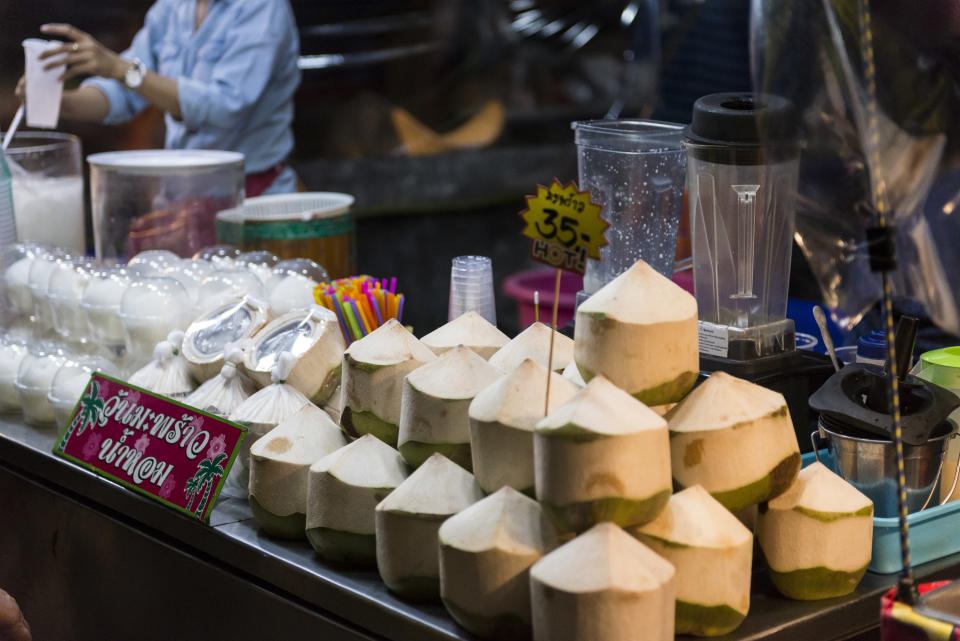 Coconuts and Thai snacks are being sold at a street stall in Tak, Thailand. The coconuts have been sliced so buyers can easily drink from them. The coconuts are a mostly white in color with a bit of green toward the bottom