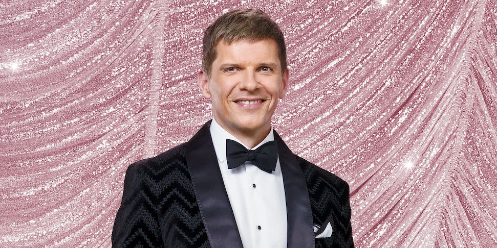 nigel harman for strictly come dancing 2023, a man stands with hands in pockets looking at the camera and smiling, he wears a black suit with bow tie and is surrounded by glitterballs