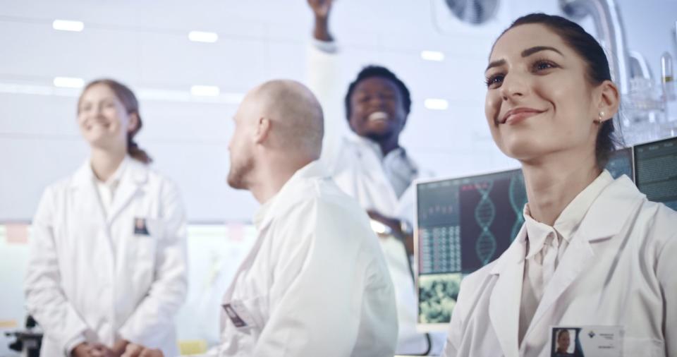 A team of scientists in a lab smile as they look at something in the distance. 