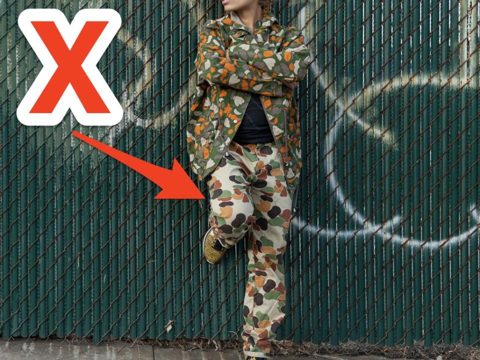 red x and arrow pointing at a woman leaning on a fence wearing camo print pants and a camo print jacket