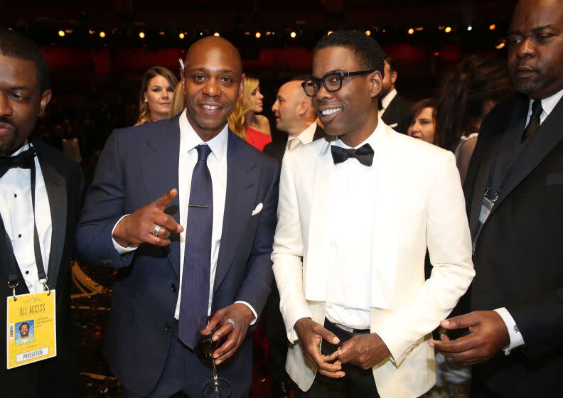 A man in a blue suit stands next to a man in a white suit and black bow tie and the two smile.