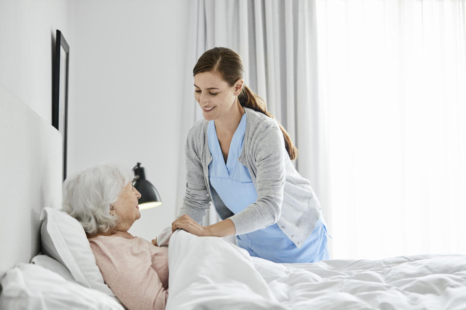Smiling caregiver putting blanket on senior woman. Confident nurse is taking care of elderly female. They are in bedroom.