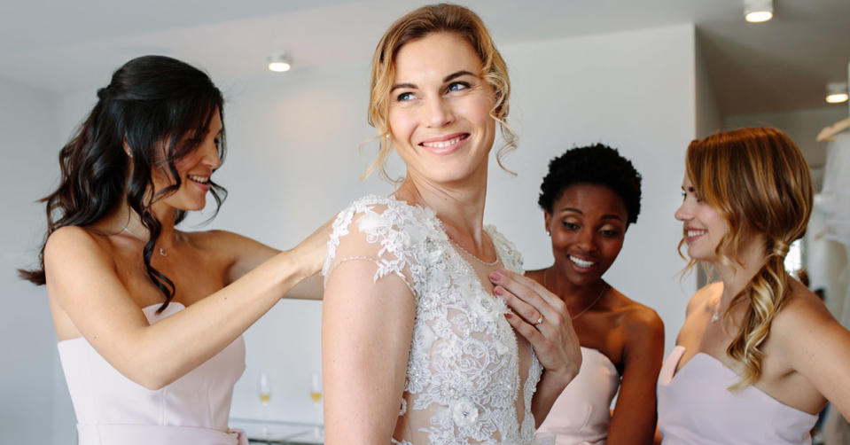 A bride in a white lace dress is smiling as 3 bridesmaids in pale pink strapless dresses tend to her