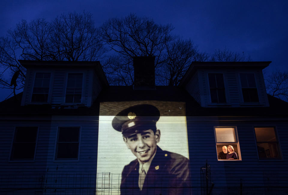 An image of veteran Emilio DiPalma, is projected onto the home of his daughter, Emily Aho, left, as she looks out a window with her husband, George, in Jaffrey, N.H., Thursday, April 30, 2020. DiPalma, a U.S. Army WWII veteran and resident of the Soldier's Home in Holyoke, Mass., died from the COVID-19 virus at the age of 93. Seeking to capture moments of private mourning at a time of global isolation, the photographer used a projector to cast large images of veterans on to the homes as their loved ones are struggling to honor them during a lockdown that has sidelined many funeral traditions. (AP Photo/David Goldman)