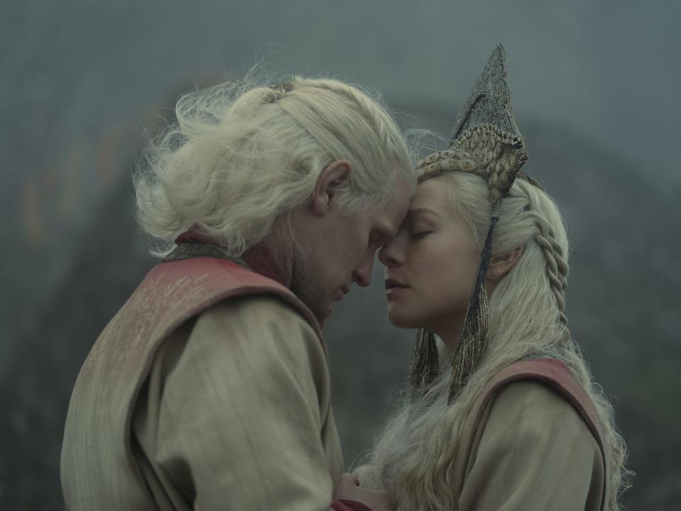 matt smith and emma d'arcy as daemon and rhaenyra targaryen, dressed in beige and red wedding garb and pressing their foreheads together. rhaneyra has a heacpiece on her head