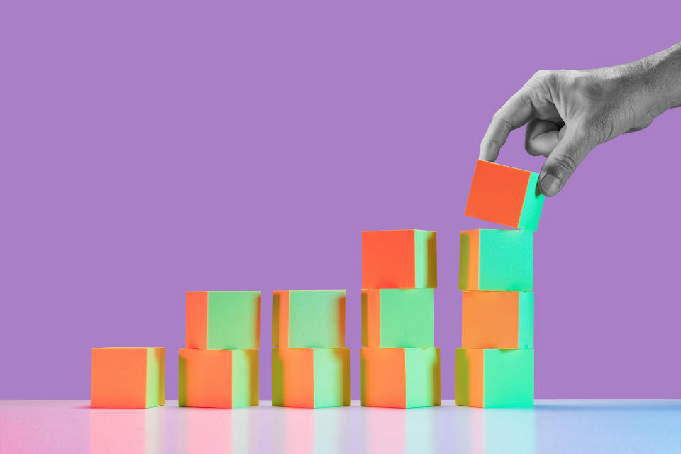 Man's hand in black and white placing block on largest stack of 5 growing stacks of yellow and red tinted blocks