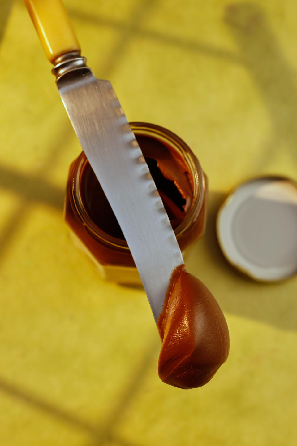 Chocolate spread on spreading knife. Knife is on top of jar. Yellowish background. Light effect.