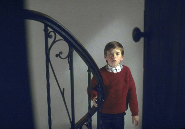 Haley Joel Osment stands on a staircase with an iron railing, looking up with a serious expression in The Sixth Sense