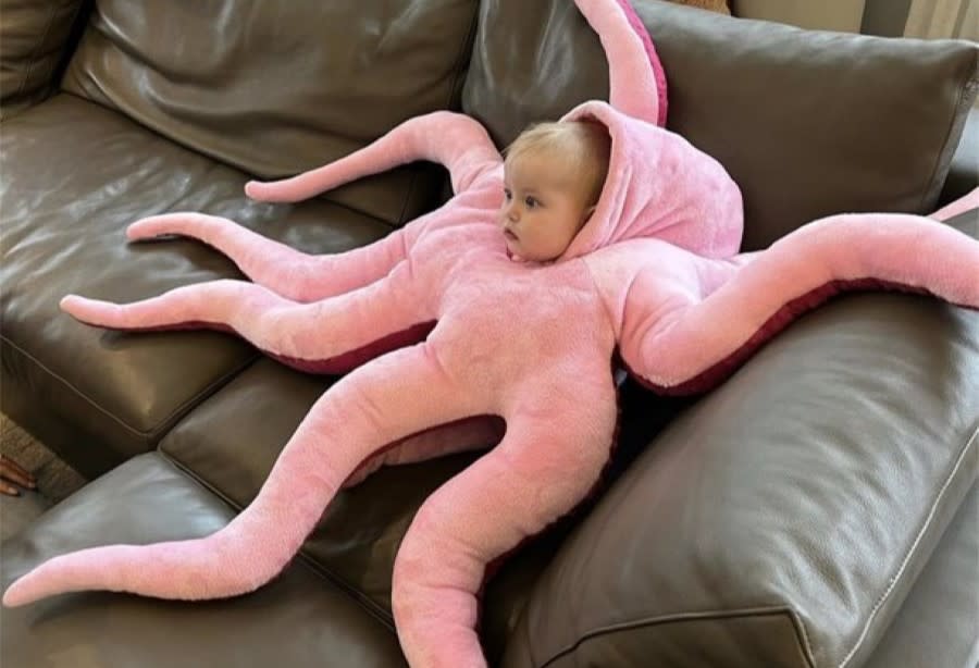 A baby sits on a couch wearing a eight-legged pink octopus costume