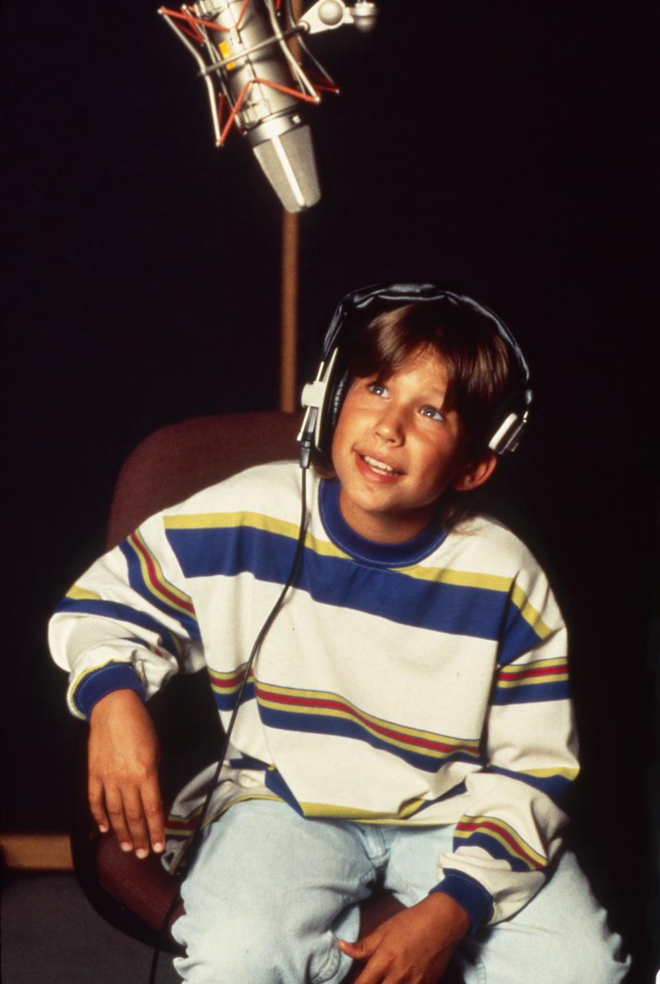 A young Jonathan Taylor Thomas wearing headphones, seated in front of a microphone in a recording studio