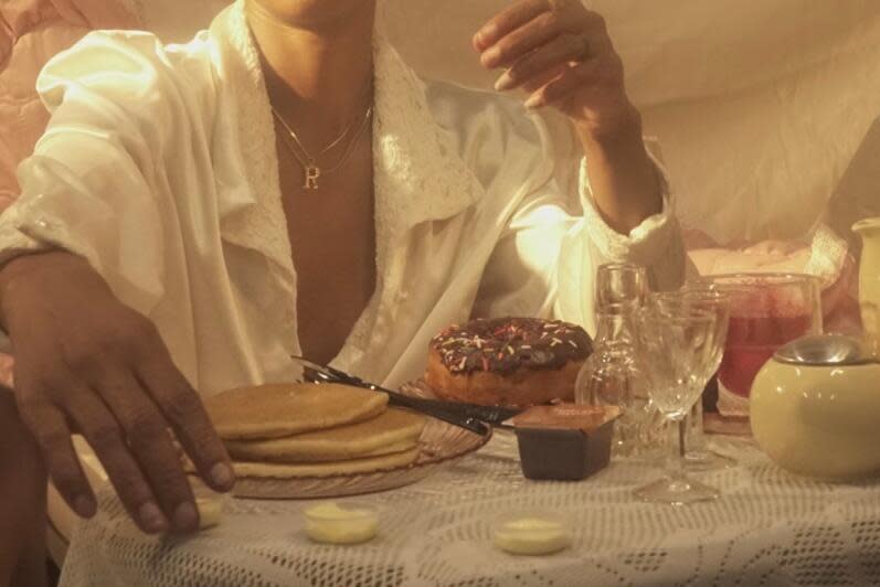 A woman in a robe, seen from neck down, sits at a table. In front of her are pancakes, chocolate doughnuts and glassware.