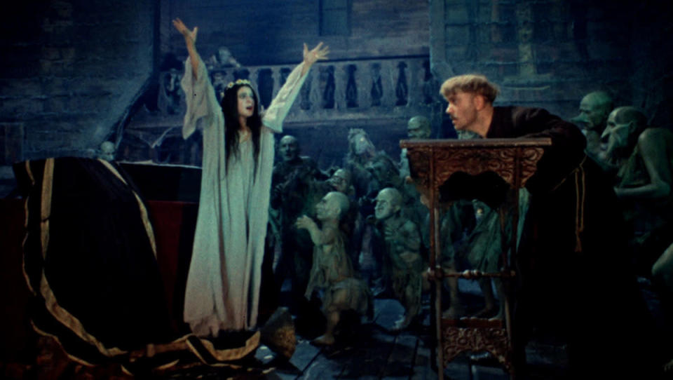A women raises her arms in front of a man bent over a table in the movie Viy.