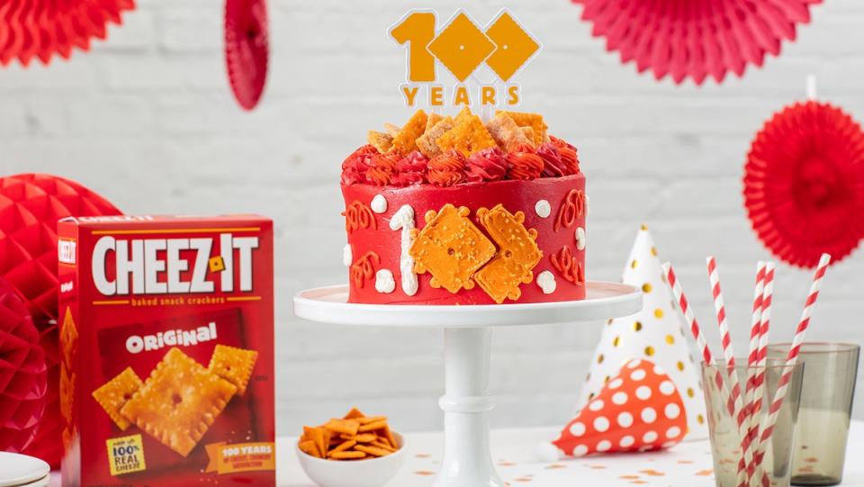 A red Cheez-It cake on a pedestal with a 100 sign sticking out of the top, all on a table with a box and bowl of Cheez-Its below it