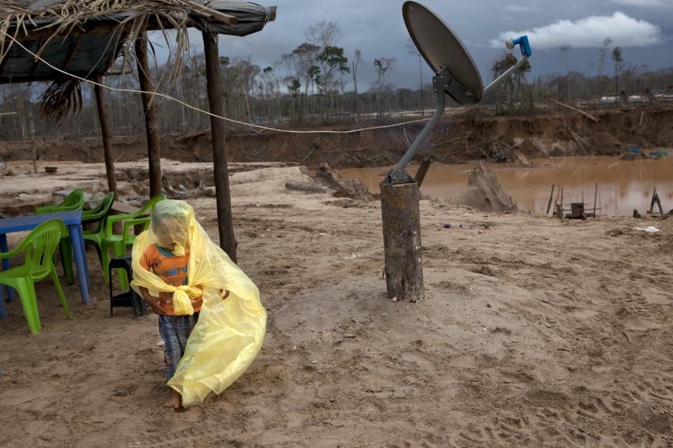 In this May 5, 2014 photo, after it stopped raining, Johan tied his father's raincoat around himself while playing in the front yard of their temporary home next to their satellite tv dish at a mining camp in La Pampa in Peru's Madre de Dios region. It's not just miners who are threatened with economic catastrophe from the government's campaign to wipe out illegal mining operations, said a mining camp cook. For every miner there is a family that eats because he works, she said. (AP Photo/Rodrigo Abd)