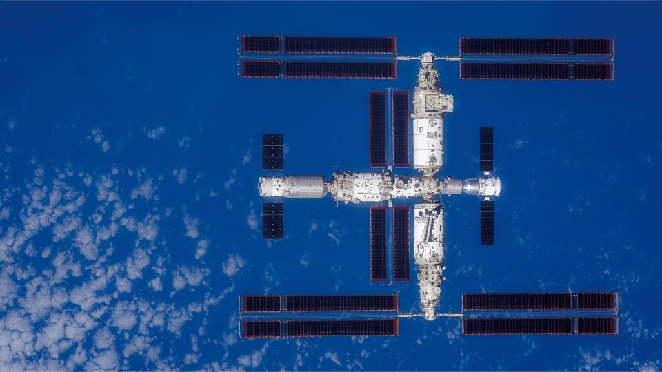 Viewed from above, a large T-shaped space station with Earth below it is visible