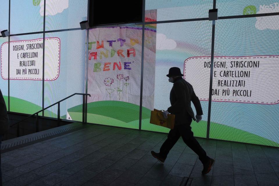 In this photo taken on Saturday, March 14, 2020 a man walks past a billboard, center, reading in Italian "Everything will be alright", in Milan, Italy. The nationwide lockdown to slow coronavirus is still early days for much of Italy, but Italians are already are showing signs of solidarity. This week, children’s drawings of rainbows are appearing all over social media as well as on balconies and windows in major cities ready, ‘’Andra’ tutto bene,’’ Italian for ‘’Everything will be alright.’’ For most people, the new coronavirus causes only mild or moderate symptoms. For some, it can cause more severe illness, especially in older adults and people with existing health problems. (AP Photo/Luca Bruno)