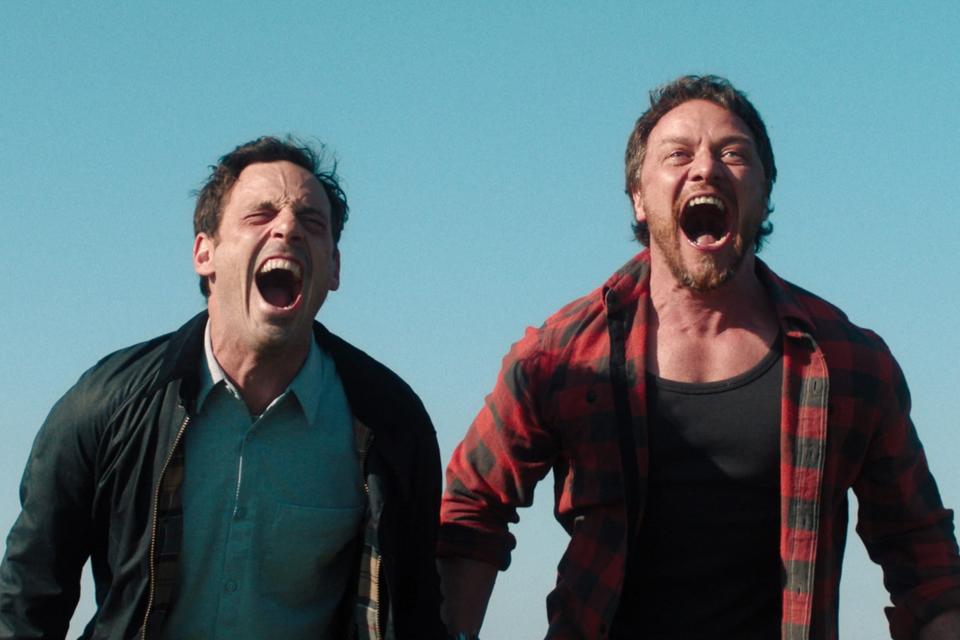 Scoot McNairy and James McAvoy are both standing outdoors, yelling with joy and excitement. Hamm wears a jacket over a shirt; McAvoy wears a plaid shirt over a tank top