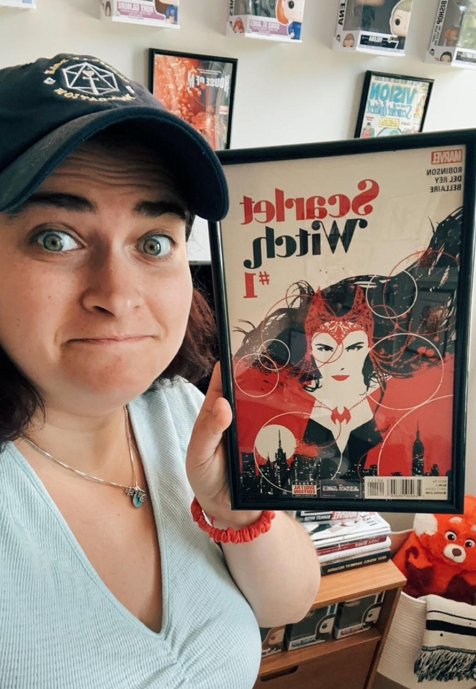 Nora, wearing a cap, holds up a framed Scarlet Witch comic book
