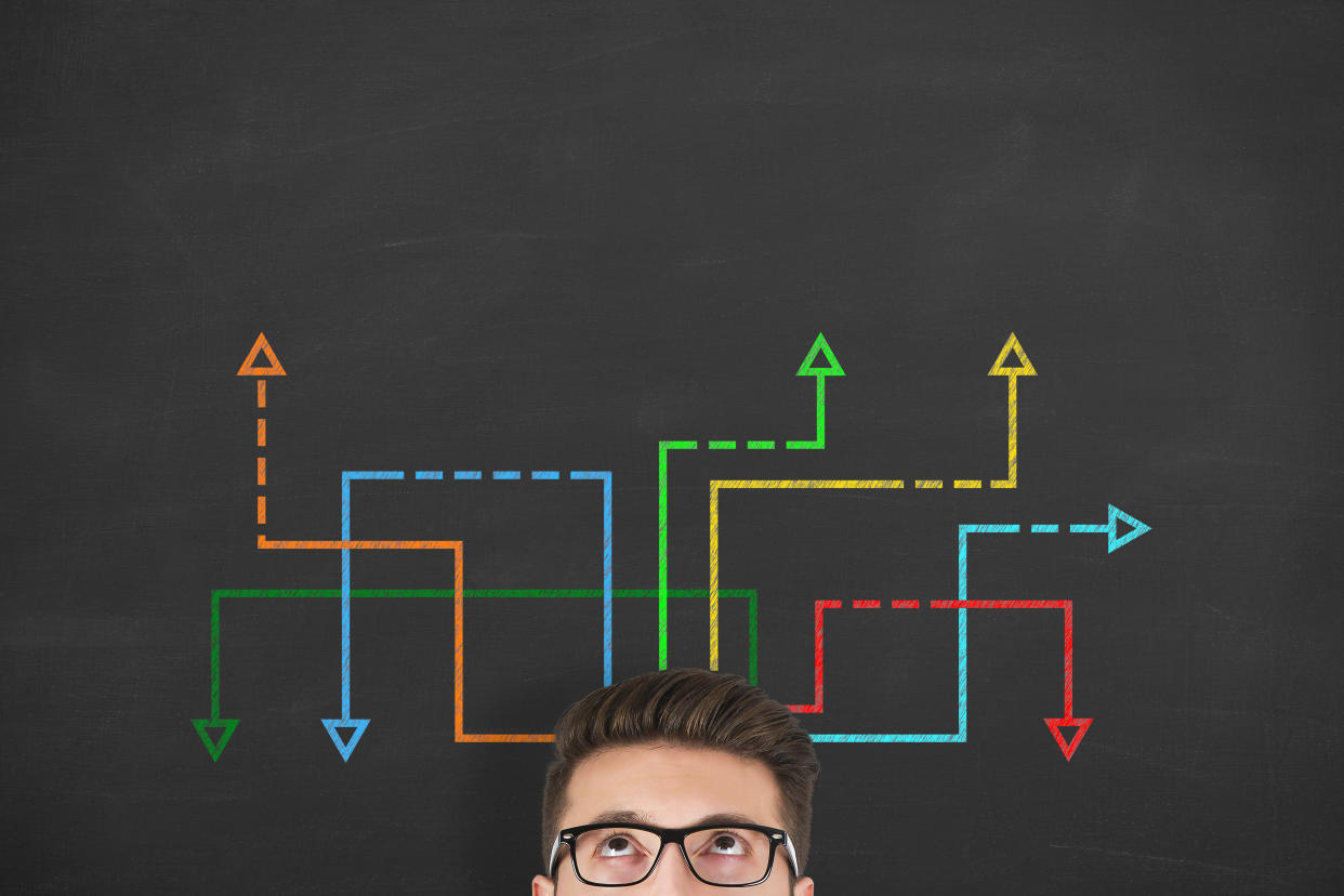 Confused, young businessman looking at many twisted arrows on the blackboard background