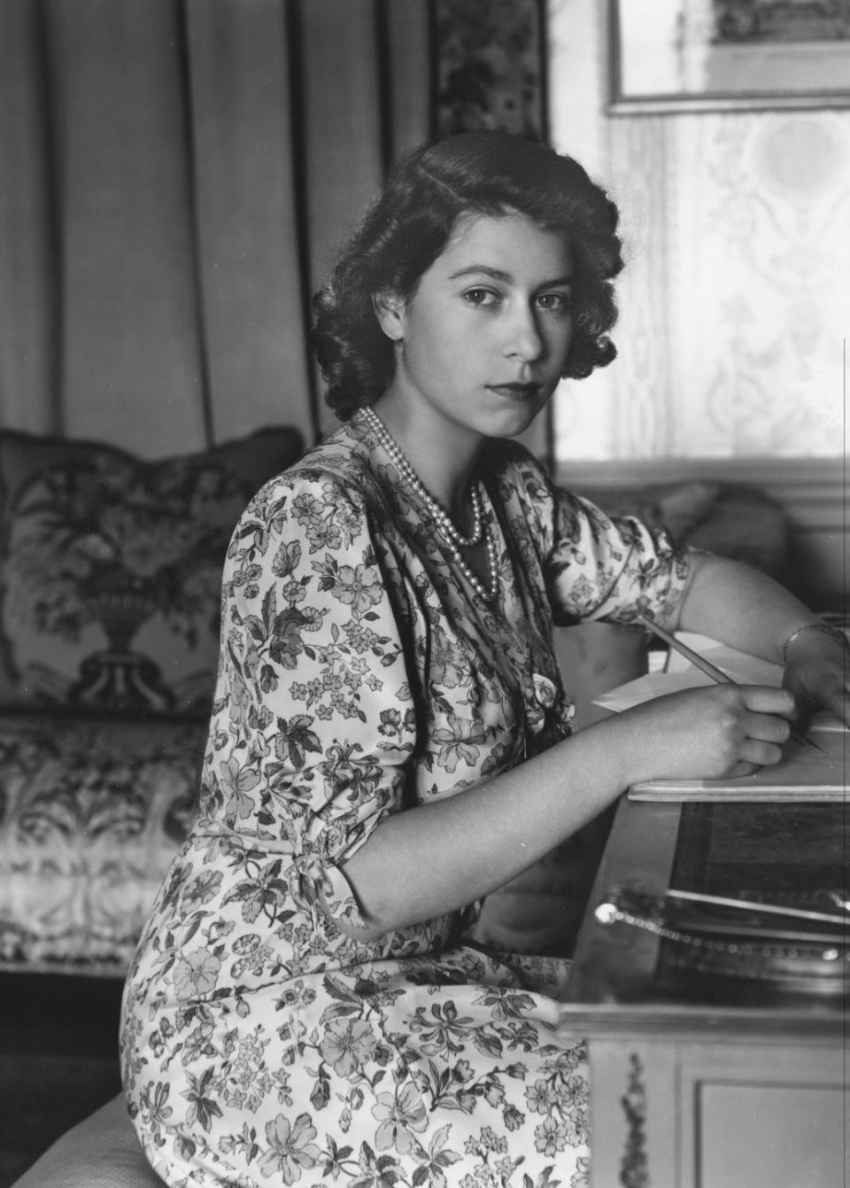 princess elizabeth looks at the camera while sitting at a desk and writing, she wears a floral dress and a double stranded pearl necklace