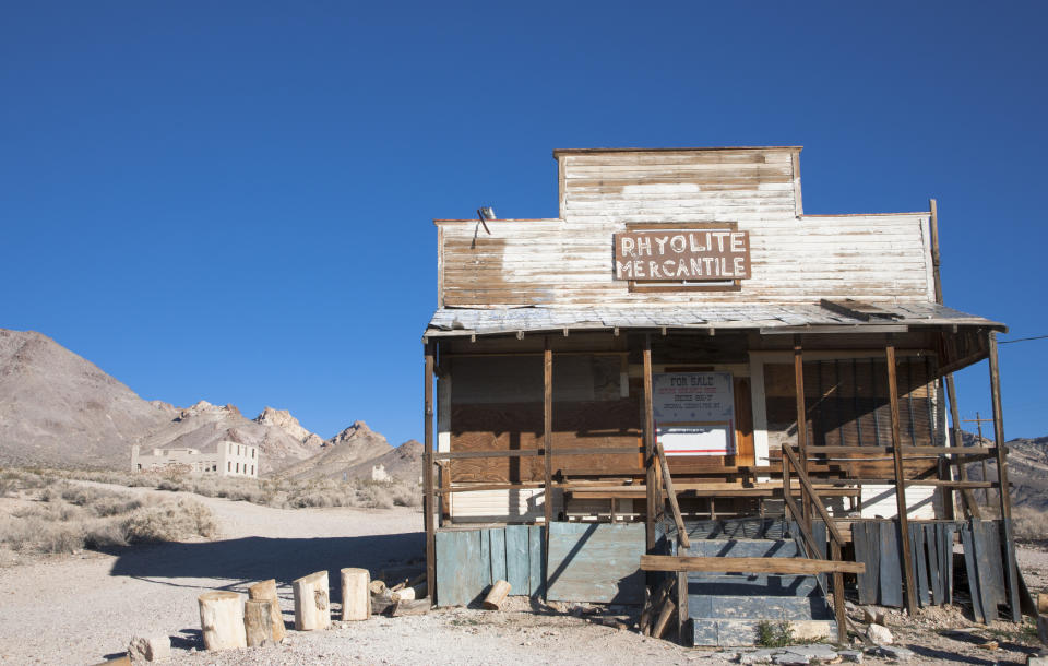 Old wooden building labeled "Rhyolite Mercantile" with abandoned structures in a desert background