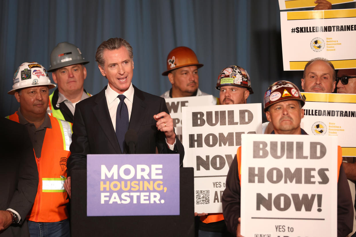 Gov. Gavin Newsom at a podium marked More Housing, Faster, surrounded by a group of men in hardhats holding signs saying: Build Homes Now! and #skilledandtrained.