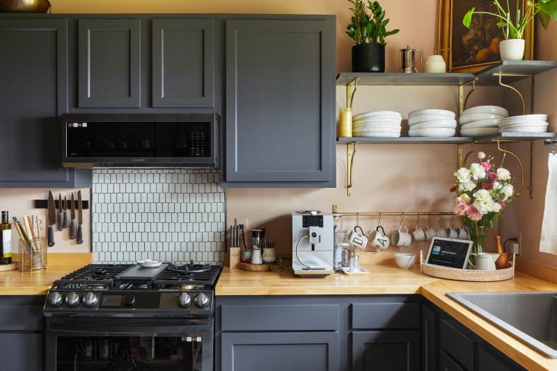 Peach kitchen with gray cabinets and shelves, wood countertops, and white backsplash with black grout over stove after renovation