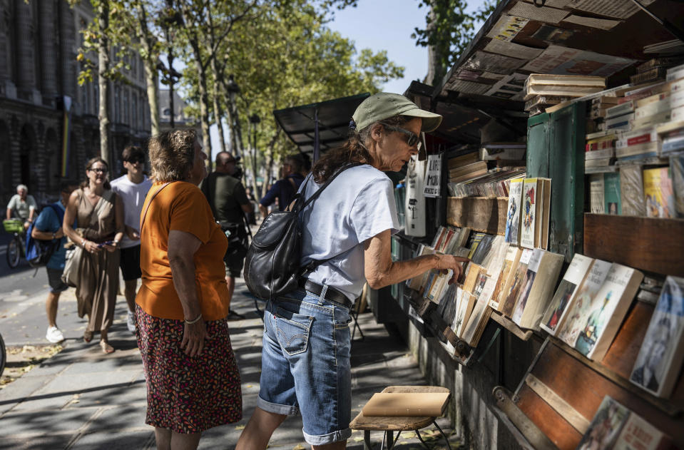 A woman looks at a bookseller booth, called "bouquiniste" along the Seine Riverbank in Paris, Tuesday, Aug. 22, 2023. The host city of Paris vowed to deliver an extraordinary grand opening on July 26, 2024, as the ceremony is expected to draw about 600,000 spectators to the Parisian quayside. Citing security measures, the Paris police prefecture ordered on July 25 the removal of 570 stationary boxes out of which booksellers have operated for decades on the quays of the Seine river. (AP Photo/Sophie Garcia)