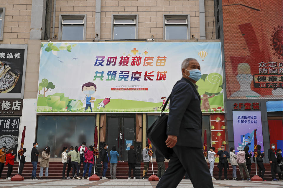 A man wearing a face mask to help curb the spread of the coronavirus walks by masked residents lining up for COVID-19 vaccine at a vaccination site with a board displaying the slogan, "Timely vaccination to build the Great Wall of Immunity together" in Beijing, Wednesday, April 21, 2021. (AP Photo/Andy Wong)