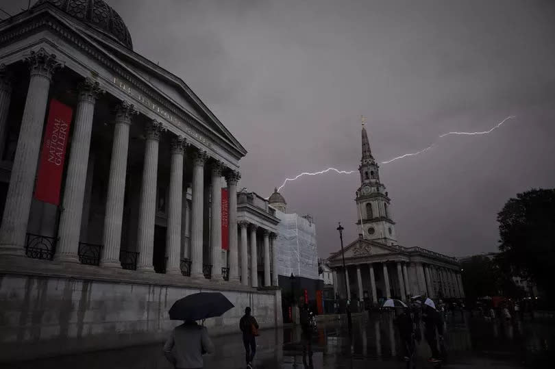 A lightning strike is pictured behind the church tower of St Martin-in-the-Fields as people walk past the National Portrait gallery under their umbrellas during a storm, in Trafalgar Square, in central London, on September 21, 2023