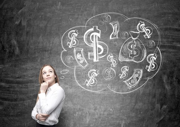 Woman with thoughtful expression looks at chalkboard with money and dollar signs drawn on it.