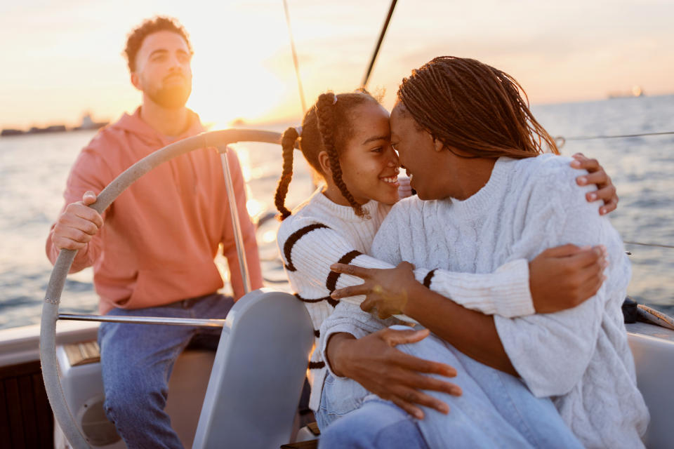 Two women embrace lovingly on a boat with a man steering in the background